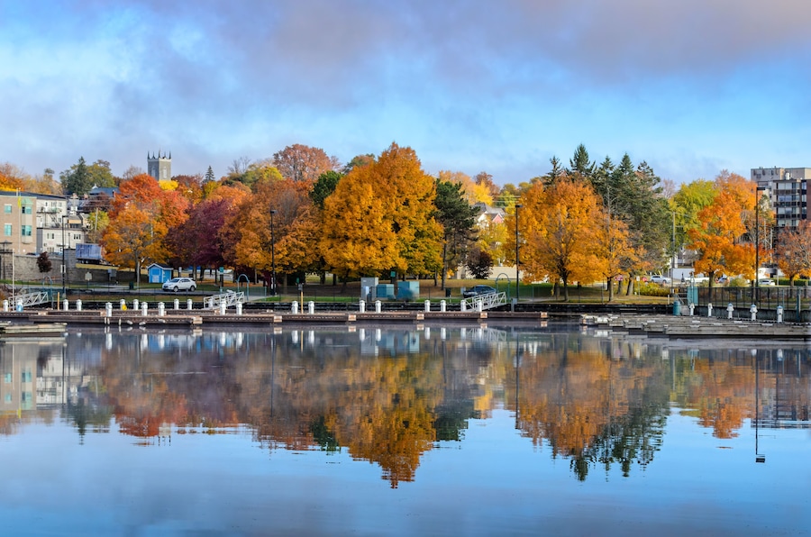 Un lago sereno que refleja árboles otoñales y el campanario de una iglesia distante.