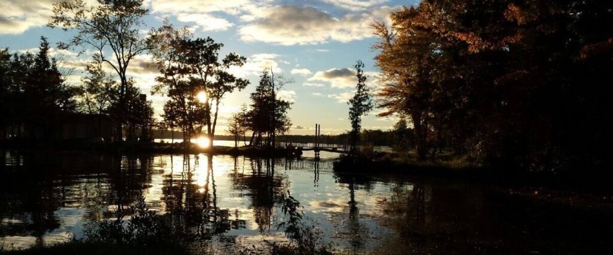 A sunset over a calm lake with trees reflecting the sky, a wooden bridge, and a distant building.