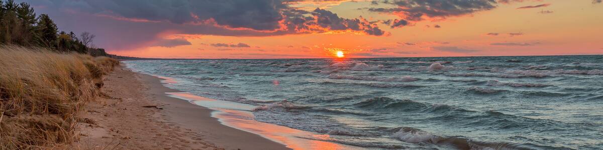 A beach with a sunset over the ocean, grassy dunes, and a clear sky.