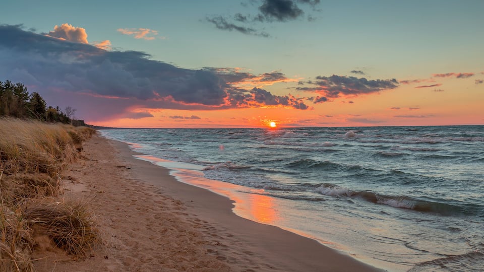 A beach with a sunset over the ocean, grassy dunes, and a clear sky.