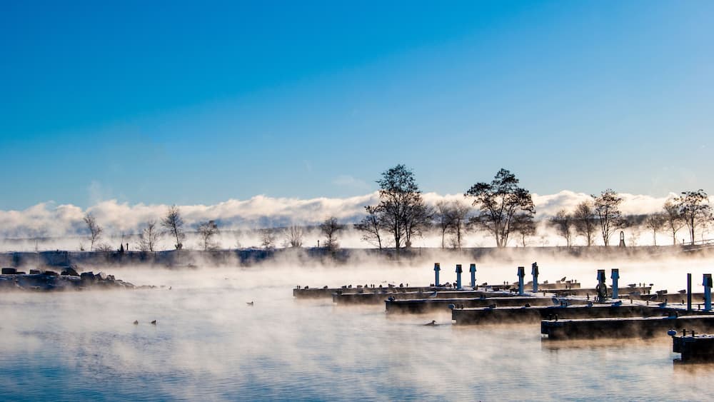 Un lac paisible, où s’élève une vapeur chaude au-dessus de l’eau, entouré d’arbres et bordé d’une rangée de bâtiments bleus.