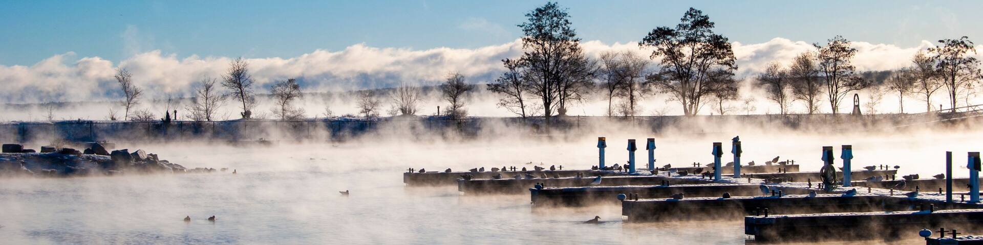 A calm lake with steam rising from the water, surrounded by trees and a row of blue structures.