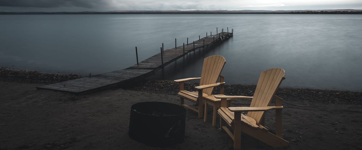 A beach with two wooden chairs and a black bucket, a wooden pier extending into calm water, and a cloudy sky.