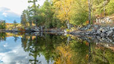 A calm lake reflecting trees with autumn foliage, surrounded by rocky terrain and dense forest.