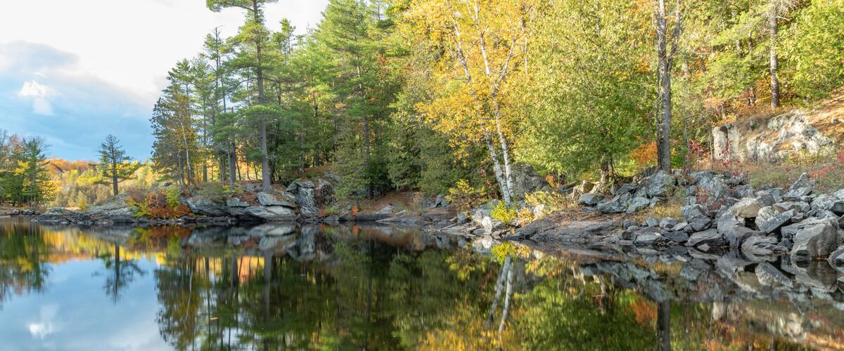 A calm lake reflecting trees with autumn foliage, surrounded by rocky terrain and dense forest.