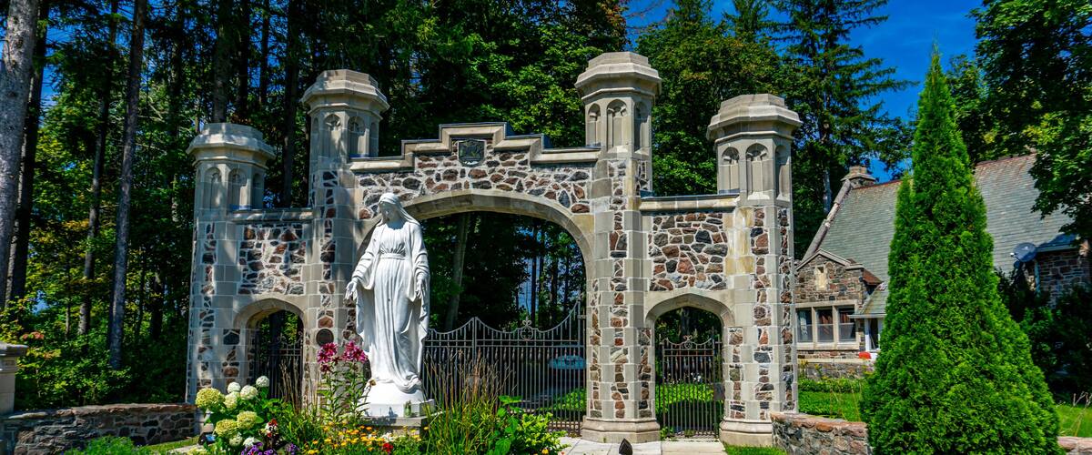 A stone archway with a statue, surrounded by a garden with flowers and a tall coniferous tree.