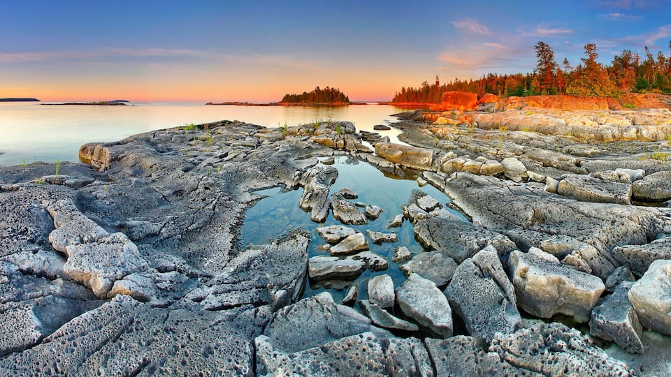 A rocky shoreline with a calm lake, a forested island, and a clear sky.