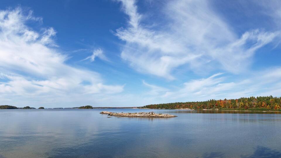 A calm lake with a small island in the center, surrounded by a forest with autumn-colored trees.