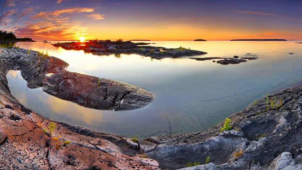 A sunset over a calm lake with rocky formations and distant islands.