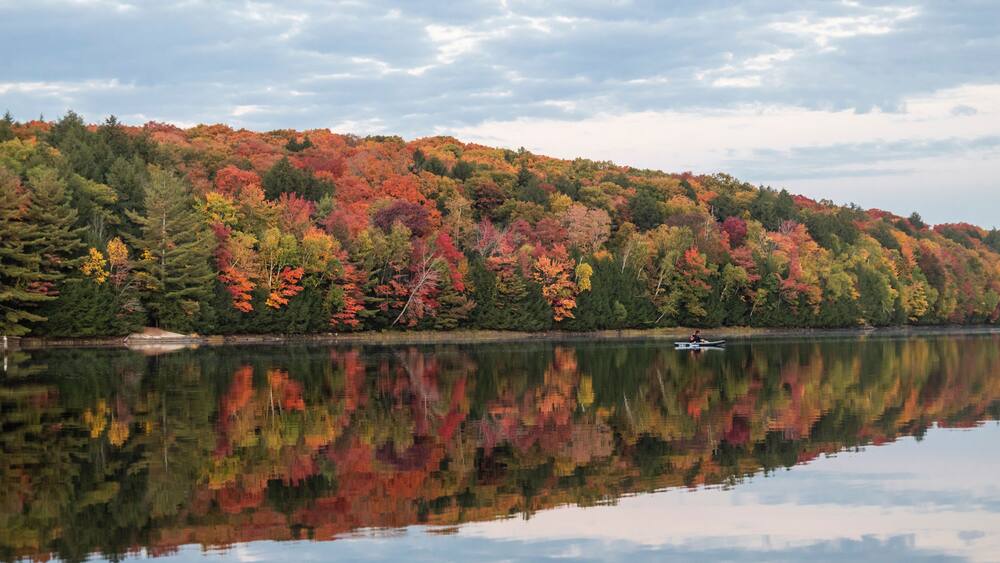 A calm lake reflecting colorful autumn foliage, a small boat on the water, and a forested hill in the background.