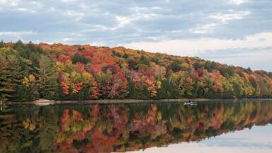 A calm lake reflecting colorful autumn foliage, a small boat on the water, and a forested hill in the background.
