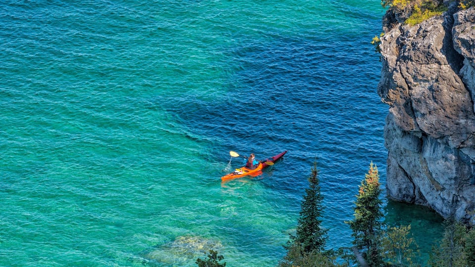 A kayaker in a turquoise sea near a rocky cliff.