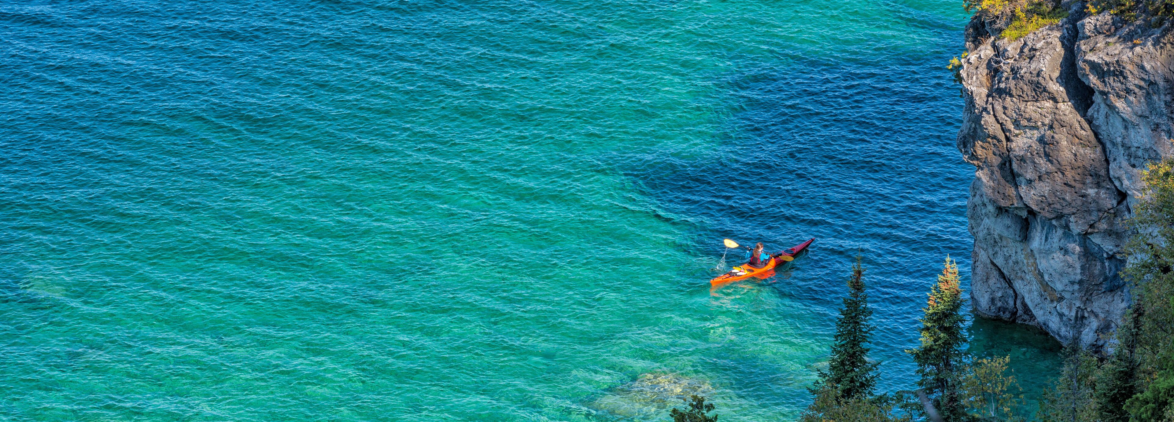 A kayaker in a turquoise sea near a rocky cliff.