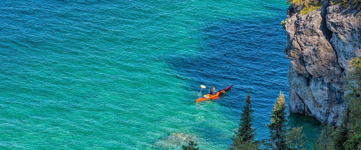 A kayaker in a turquoise sea near a rocky cliff.