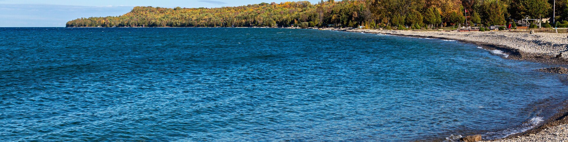 A rocky shoreline with clear blue water, a forested hill in the background, and a clear sky.