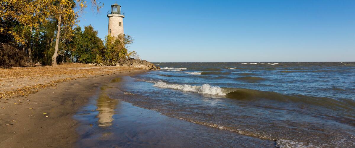 A lighthouse by a beach with waves, trees, and a clear sky.