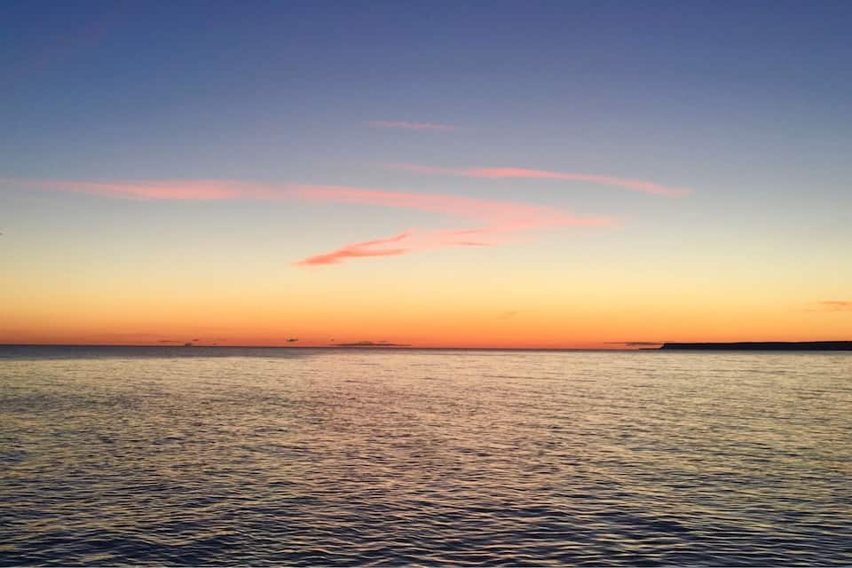 A sunset over a calm sea with a pinkish sky and distant land.