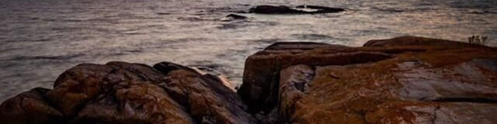 A sunset over a rocky coastline with the sun partially visible near the horizon.