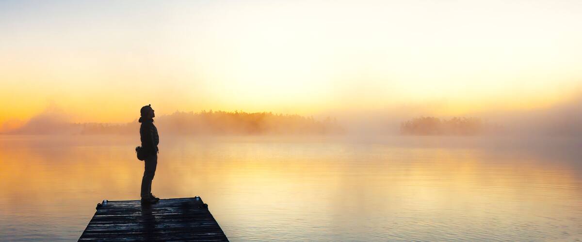 A person standing on a wooden dock at sunrise with mist over a calm body of water.