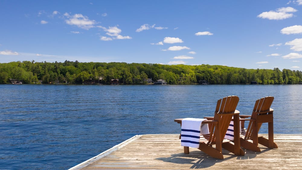 Two Adirondack chairs with neatly folded beach towels sit on a wooden dock, overlooking a calm Muskoka lake in Ontario, Canada. Across the water, trees and cottages create a peaceful background.