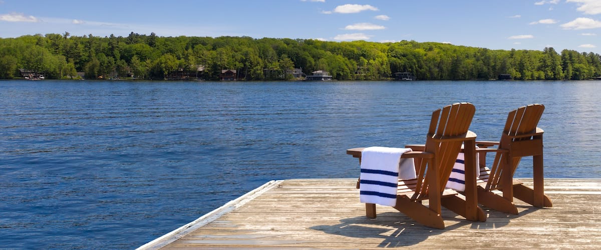 Two Adirondack chairs with neatly folded beach towels sit on a wooden dock, overlooking a calm Muskoka lake in Ontario, Canada. Across the water, trees and cottages create a peaceful background.
