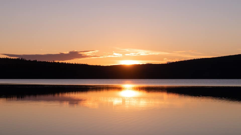 A sunset over a calm lake with a forested hill in the background.