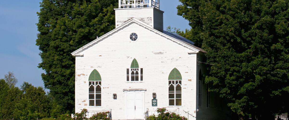 A white church with a steeple and arched windows, surrounded by trees and a grassy field.
