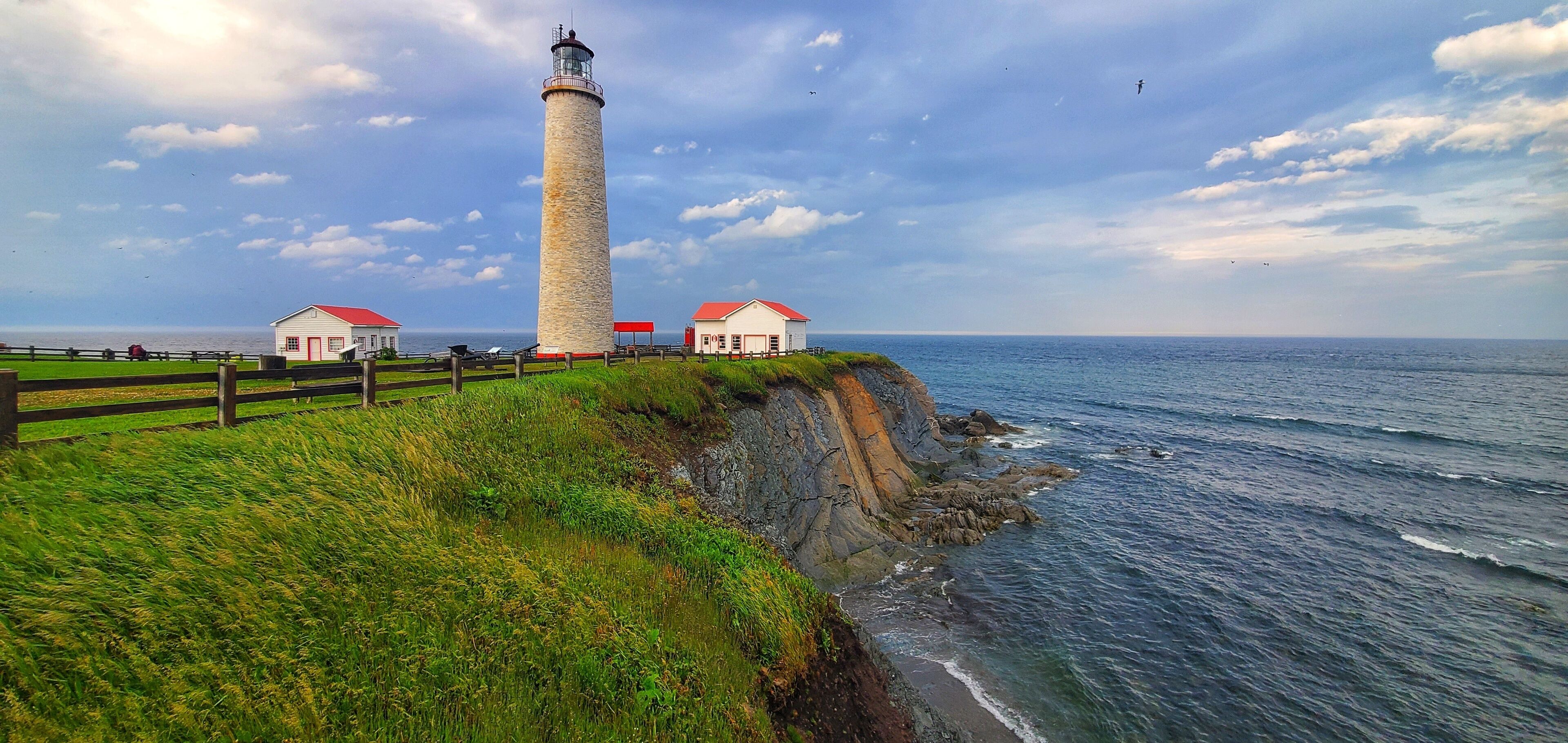 A lighthouse, grassy cliff, and two small buildings with red roofs by the ocean.