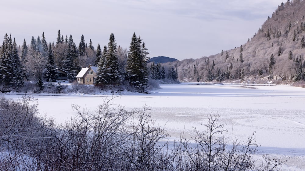 A snow-covered landscape with a house, dense evergreen trees, and a frozen lake.