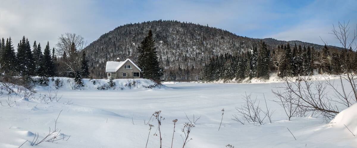 A snow-covered landscape with a wooden cabin and a forested mountain in the background.