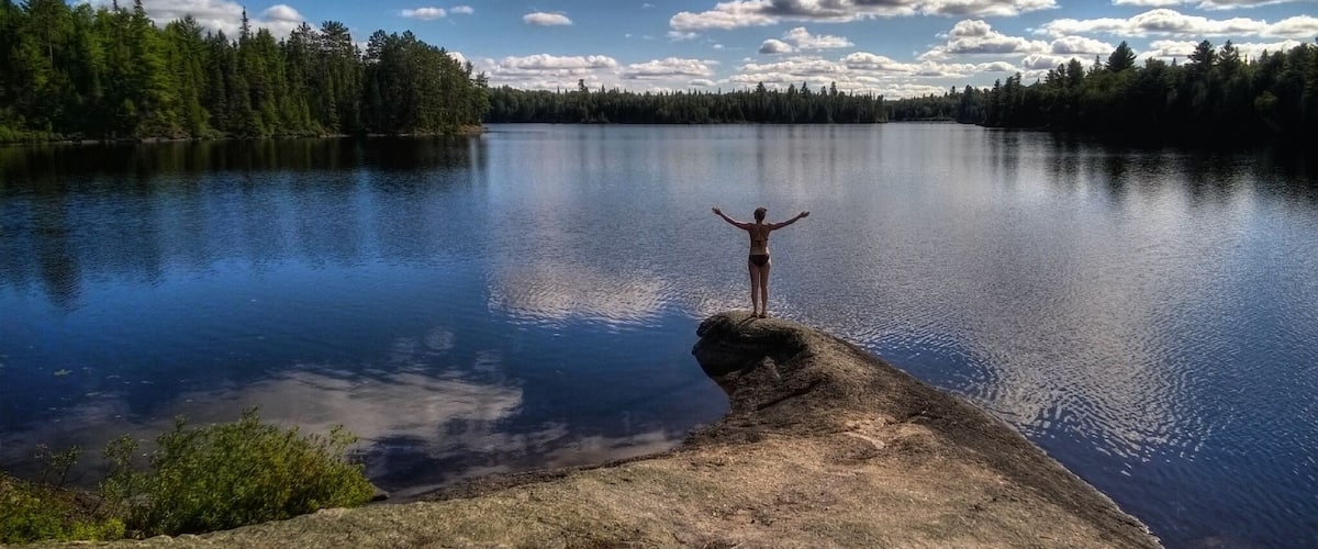 Une personne se tient sur un rocher, les bras tendus au-dessus d’un lac paisible, entouré d’une forêt dense.