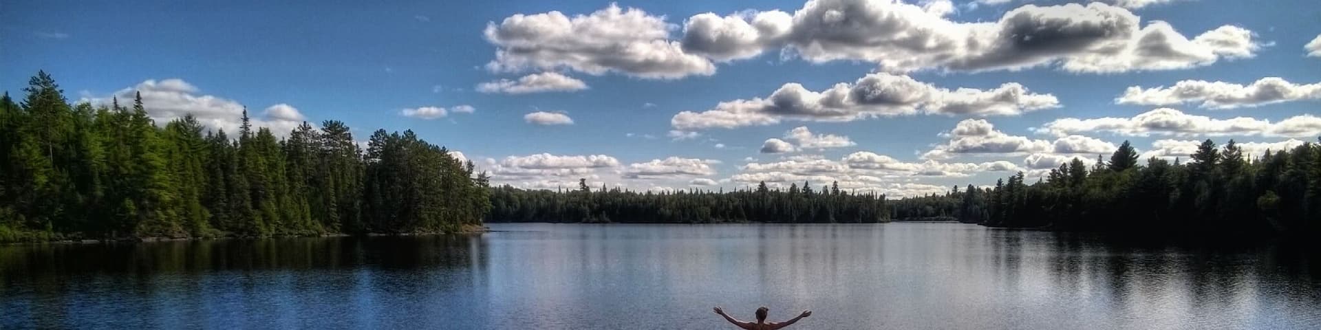 A person standing on a rock with arms outstretched over a calm lake surrounded by dense forest.