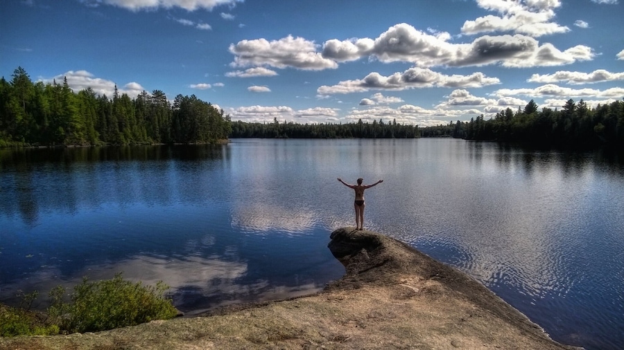 A person standing on a rock with arms outstretched over a calm lake surrounded by dense forest.