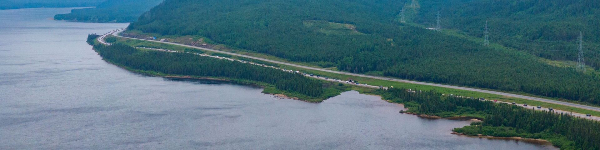 A coastal road winding through a forested valley with mountains in the background.
