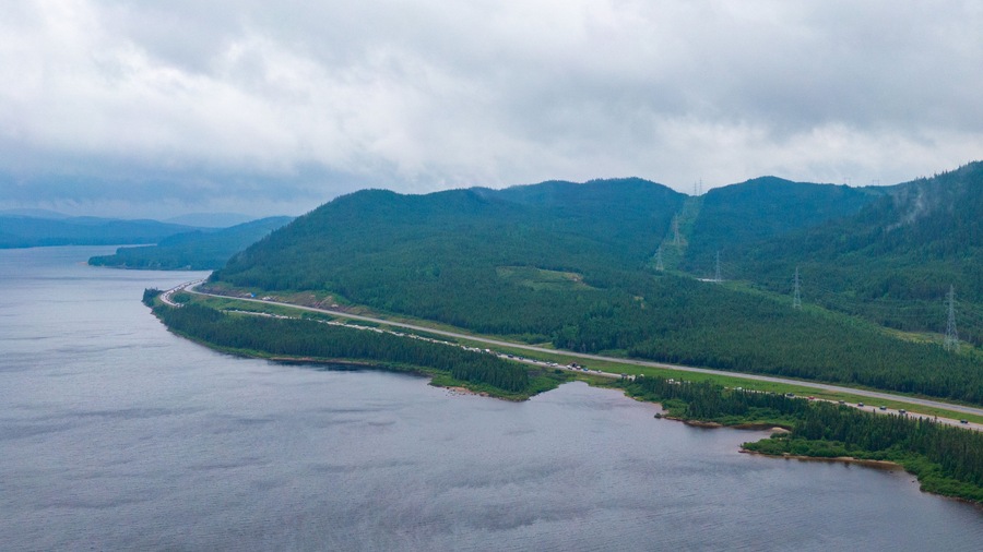 A coastal road winding through a forested valley with mountains in the background.