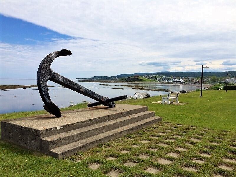 Escultura de un gran ancla sobre una plataforma de hormigón, junto a una masa de agua, con un banco y zona verde al fondo.