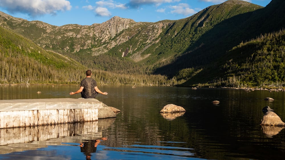 A person meditating on a concrete platform by a calm lake surrounded by mountains.