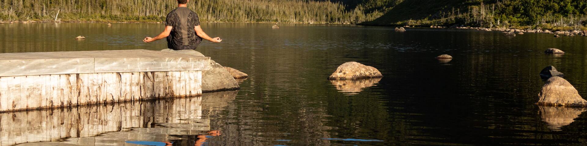 A person meditating on a concrete platform by a calm lake surrounded by mountains.