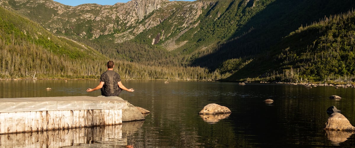 A person meditating on a concrete platform by a calm lake surrounded by mountains.