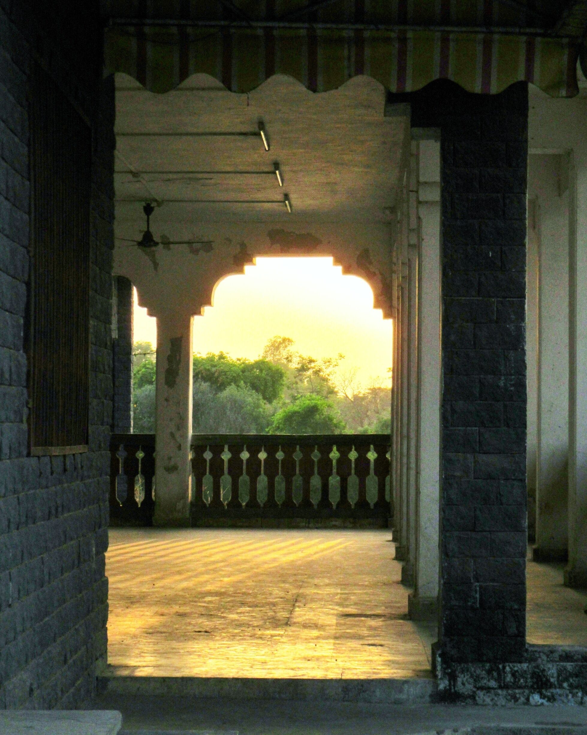 An arched corridor with a wooden floor, brick walls, and a railing overlooking greenery.