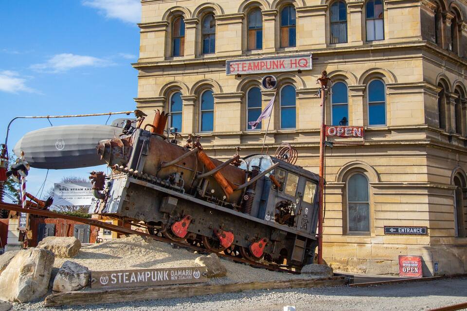 Uma locomotiva a vapor sobre trilhos, em frente a um edifício com uma placa indicando "STEAMPUNK HQ".