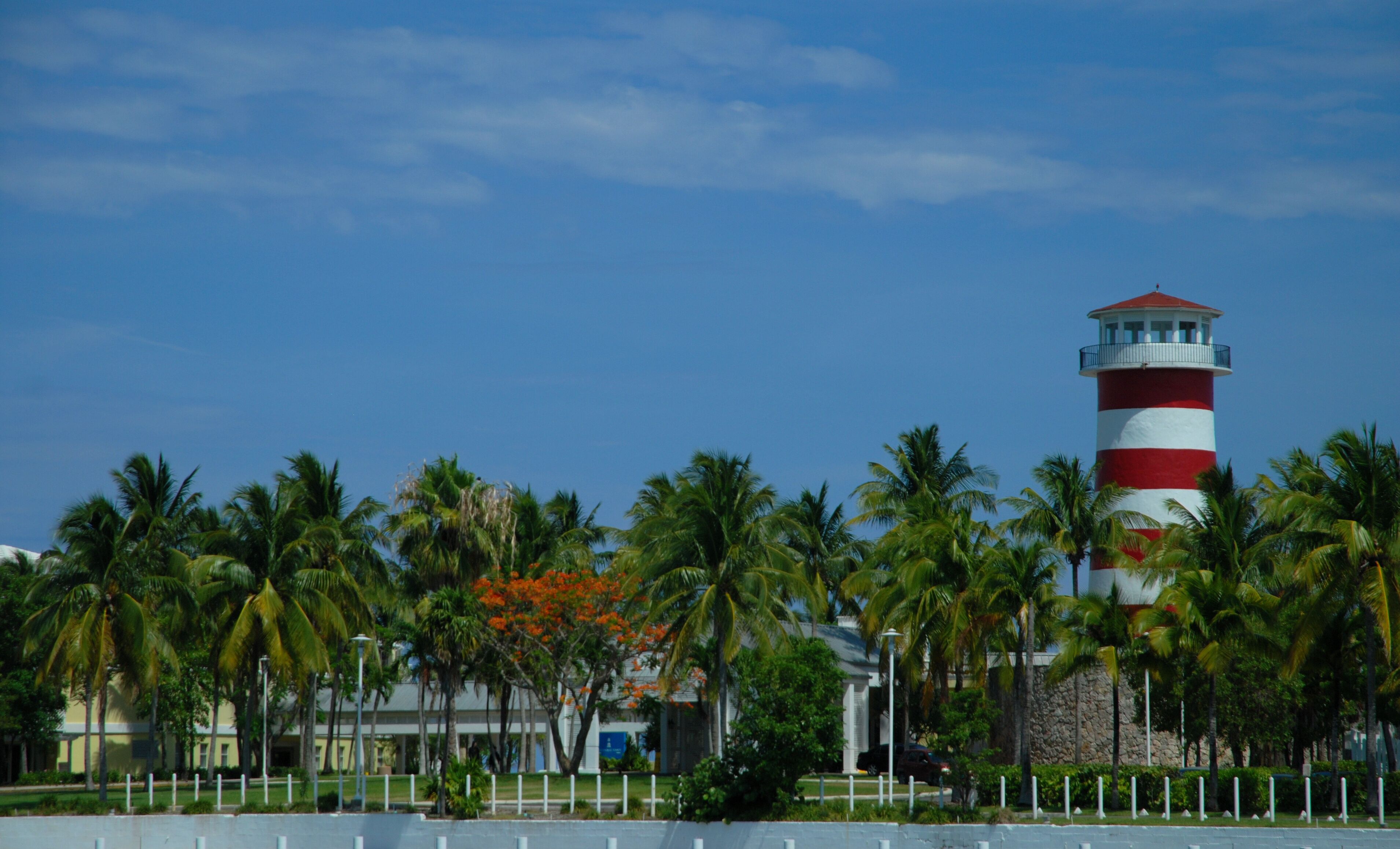 A lighthouse with red and white stripes, surrounded by palm trees and a building with a red roof.