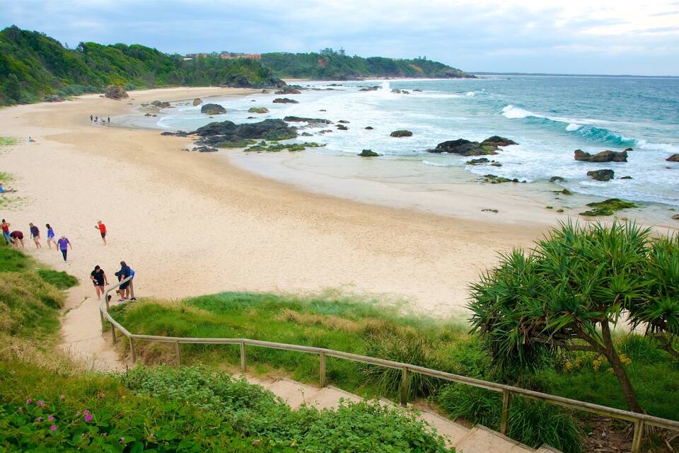 A beach with people walking along the shore, surrounded by greenery and rocky outcrops.