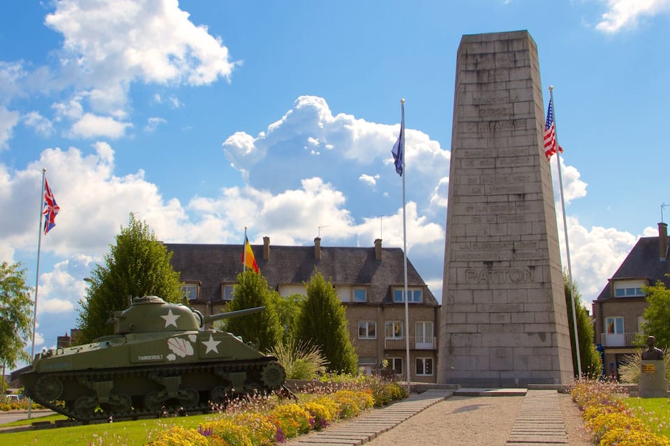 Um monumento com inscrições, um tanque de guerra e bandeiras em um jardim, com edifícios ao fundo.