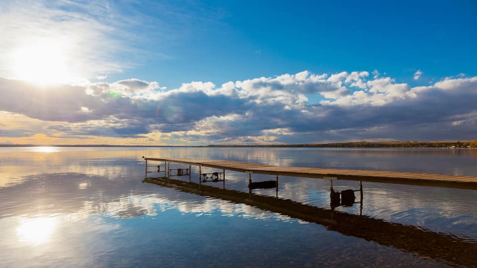 A calm lake with a wooden dock extending into the water, surrounded by a clear sky and distant trees.