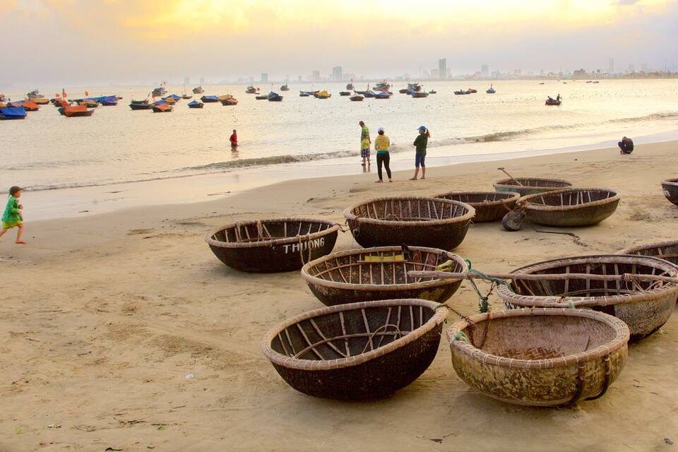A beach with several large woven baskets and boats on the sand, a few people walking, and a city skyline in the background.