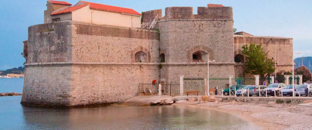 A historic stone fortress with a red-tiled roof, situated by a calm body of water.