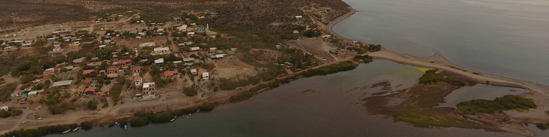 An aerial view of a coastal town with a beach, houses, and a mountainous backdrop.
