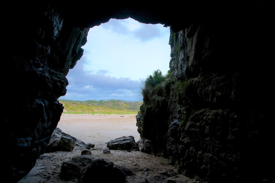 A cave entrance leading to a beach with a cloudy sky.
