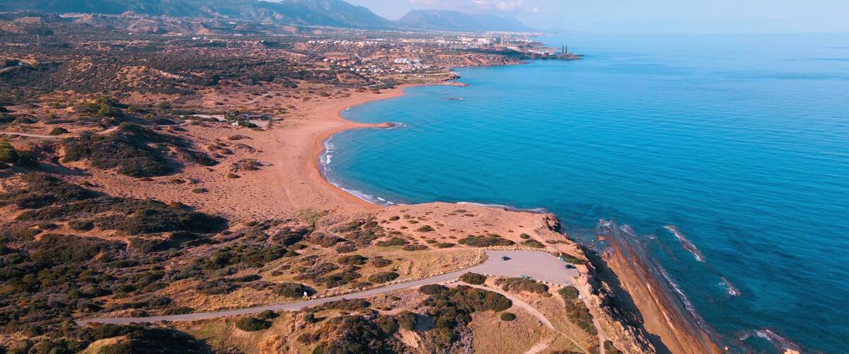 An aerial view of a coastal area with a sandy beach, a winding road, and a mountainous backdrop.